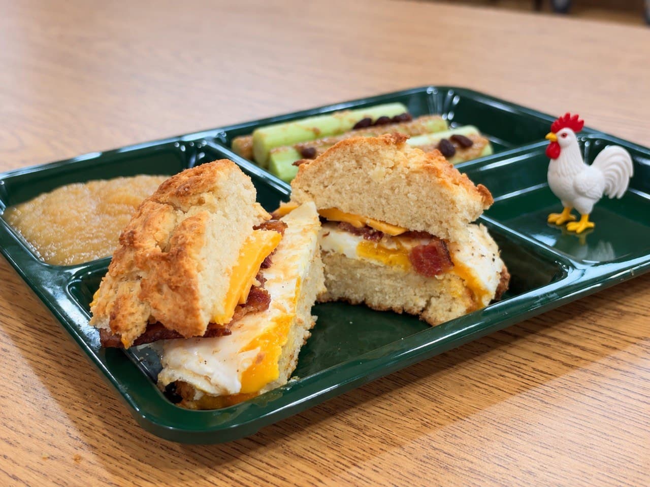 A large drop biscuit breakfast sandwich split to show egg, bacon and melted cheese, on a green lunch tray beside applesauce and celery.
