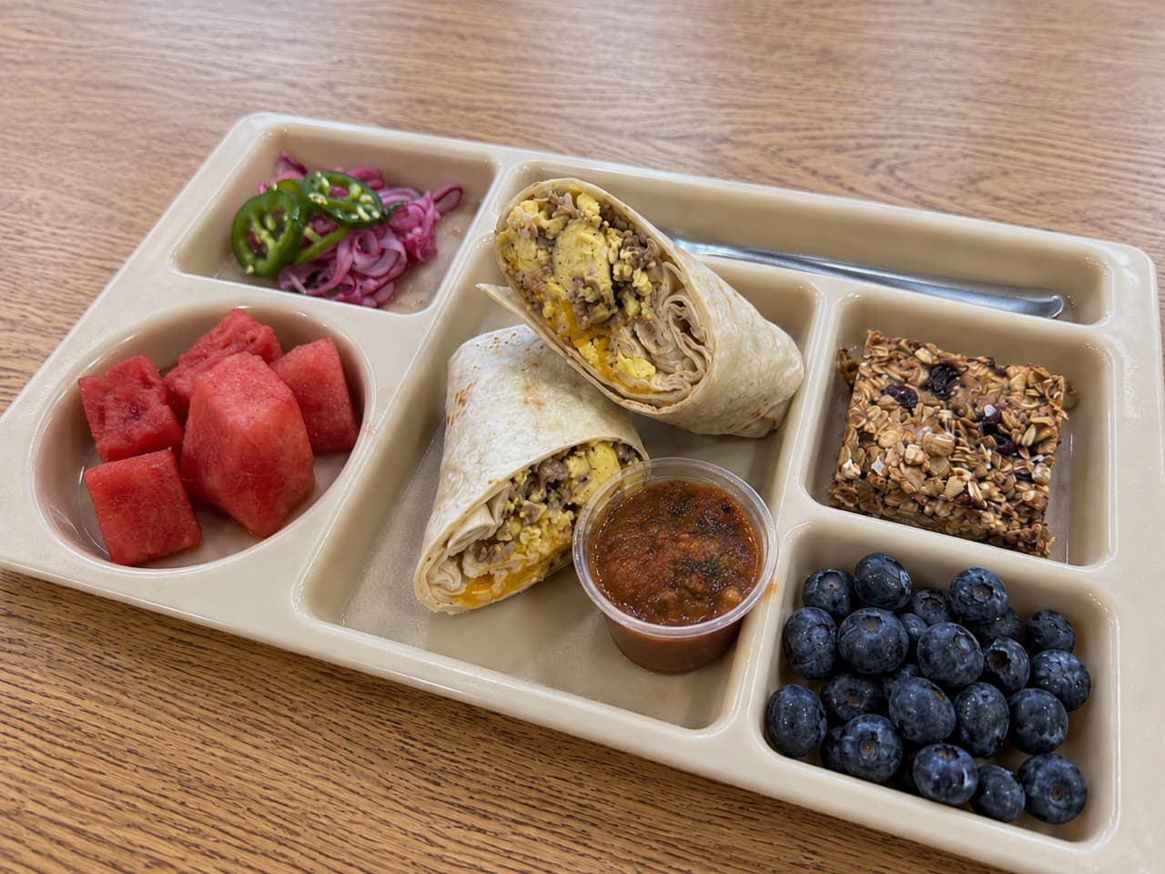 Breakfast burrito halves with egg, sausage and cheese, beside salsa, a granola square, watermelon, blueberries and pickled vegetables.