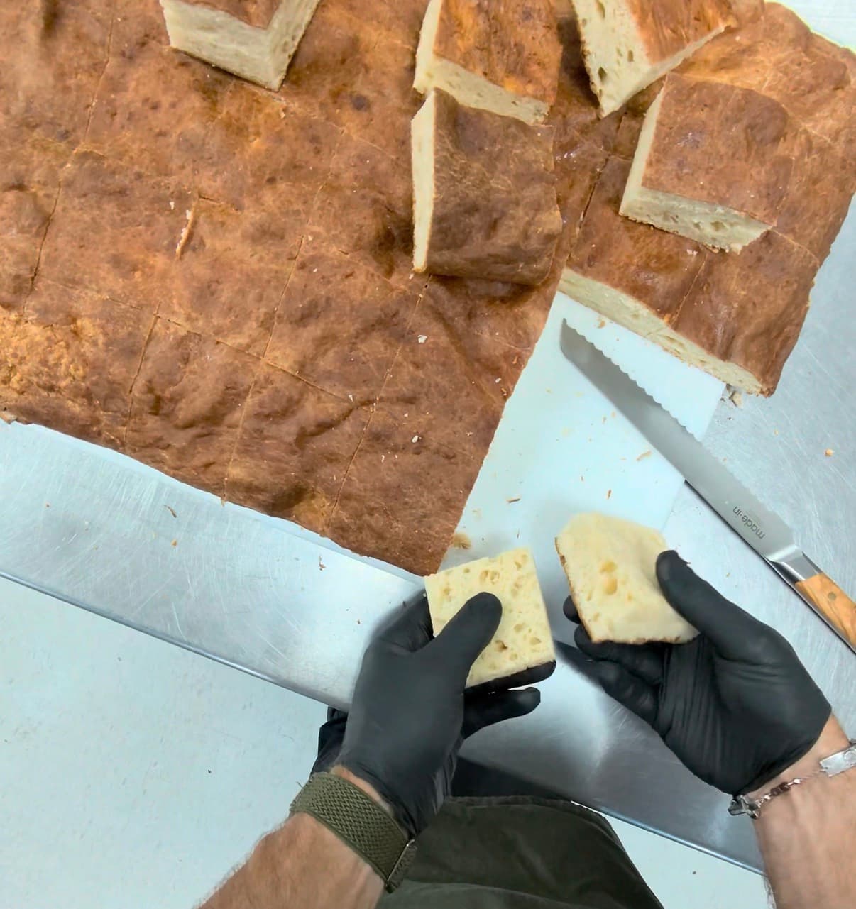 Fresh-baked flatbread cut into squares, with gloved hands holding a split piece to show the crumb.