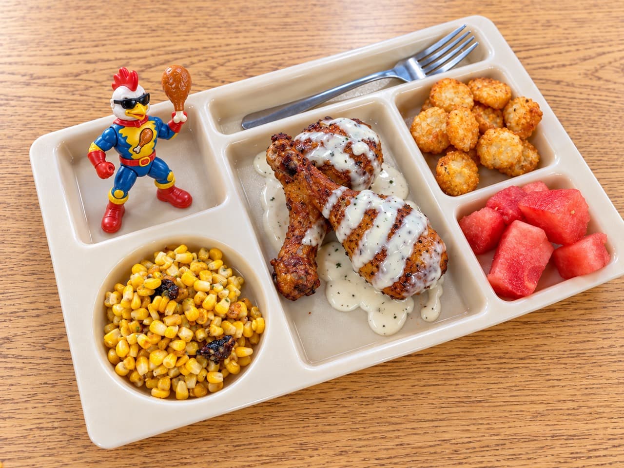 Roasted chicken drumsticks with Golden Ranch on a lunch tray beside corn, tater tots and watermelon.