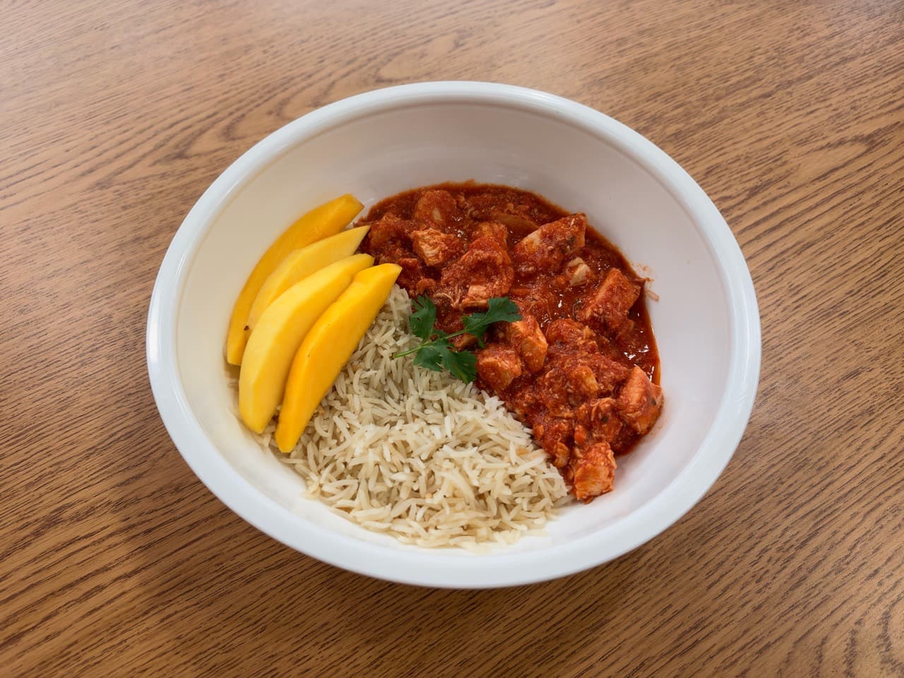 Chicken tikka masala with basmati rice, sliced mango and cilantro in a white bowl.