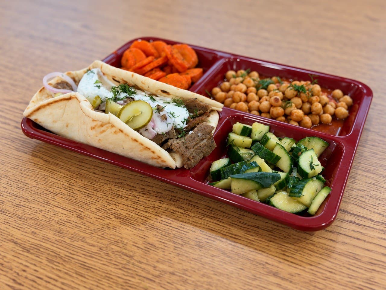 Gyro meat in a pita with tzatziki on a red lunch tray beside honey dill carrots, stewed tomato chickpeas and cucumber salad.