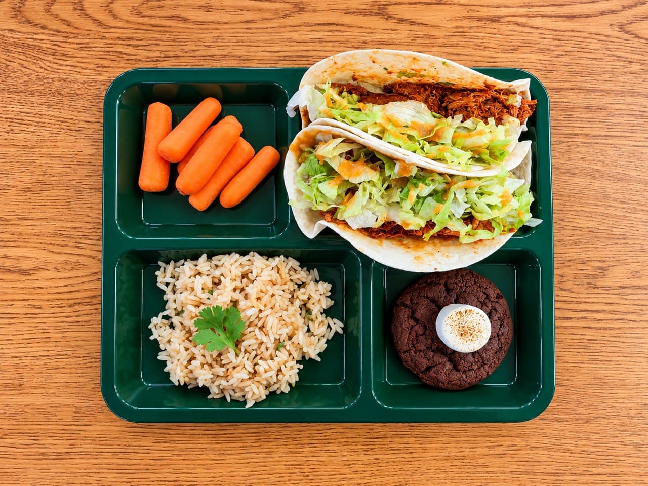 Shredded guajillo chicken tacos with lettuce and sauce on a green tray beside carrots, rice and a marshmallow-topped chocolate cookie.