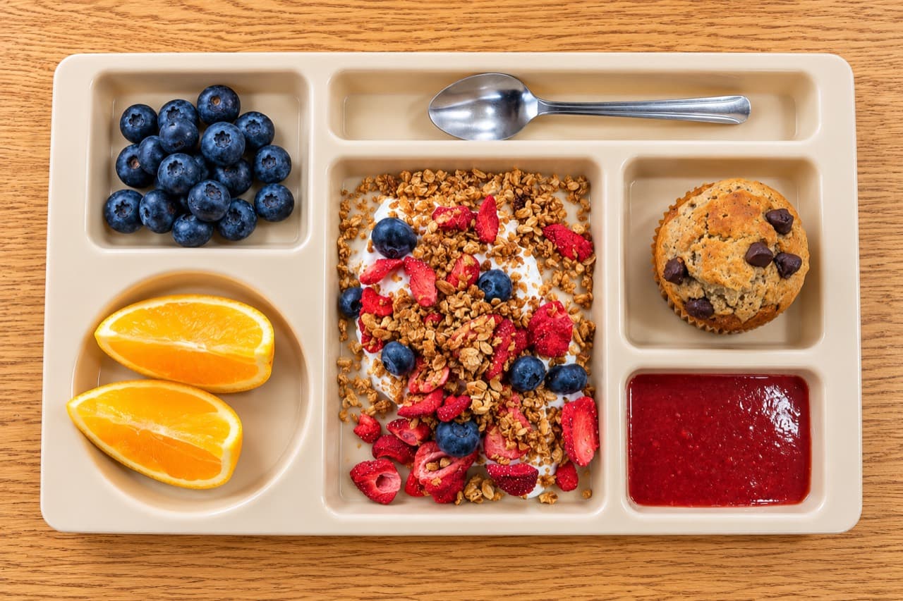 Greek yogurt with toasted granola and berries, beside a chocolate chip pancake muffin and strawberry jam.