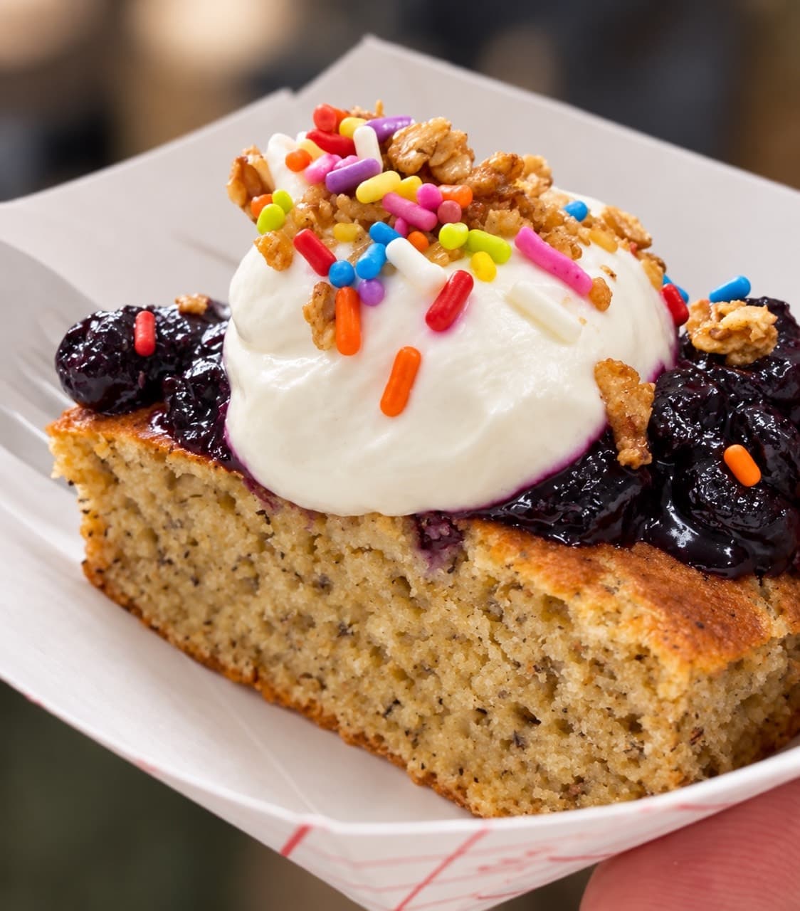 A square of pancake bake topped with blueberry compote, whipped yogurt cream, granola and rainbow sprinkles.