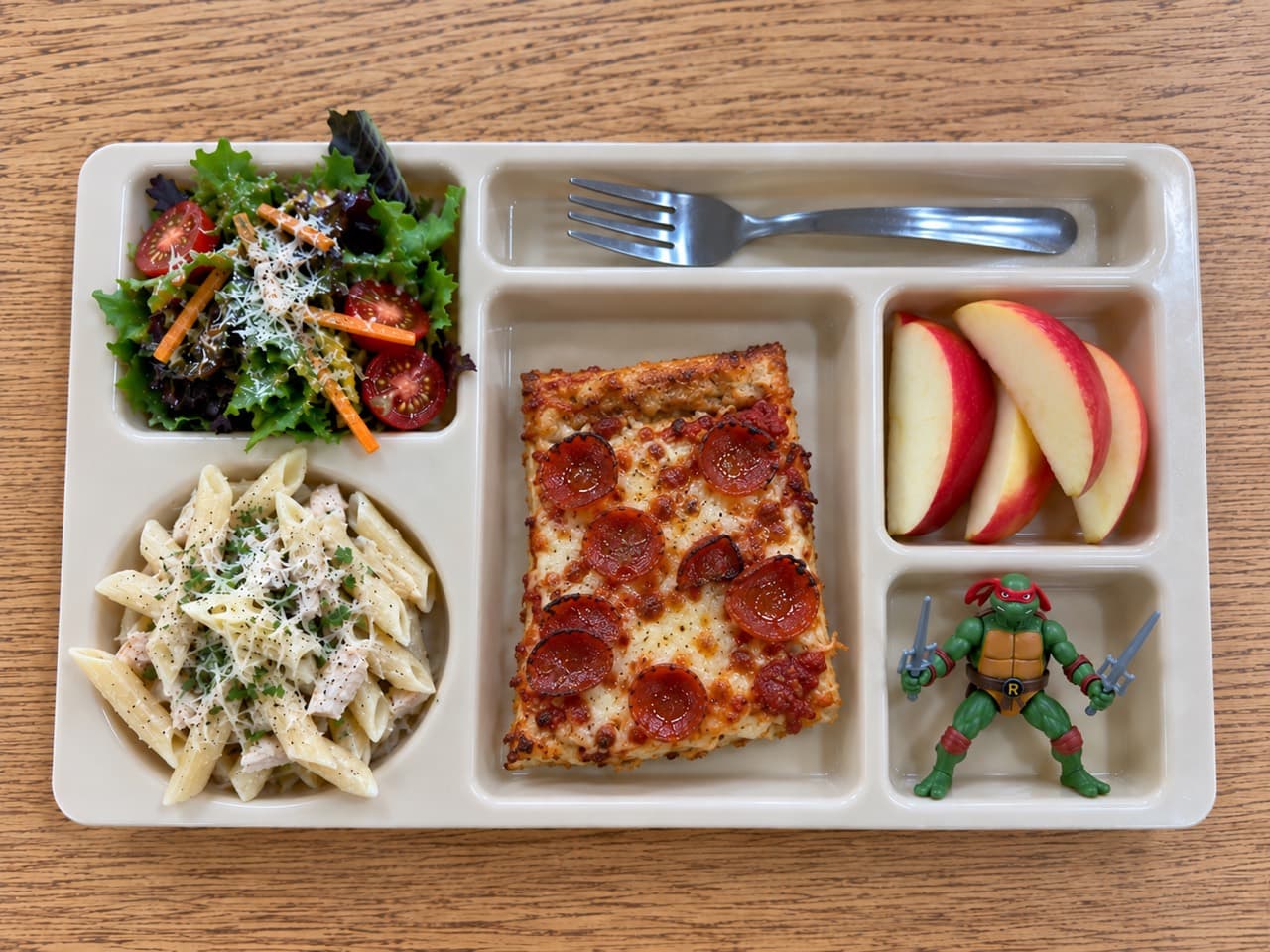A rectangular pepperoni pizza slice on a lunch tray with green salad, pasta salad, apple slices and a small toy turtle.