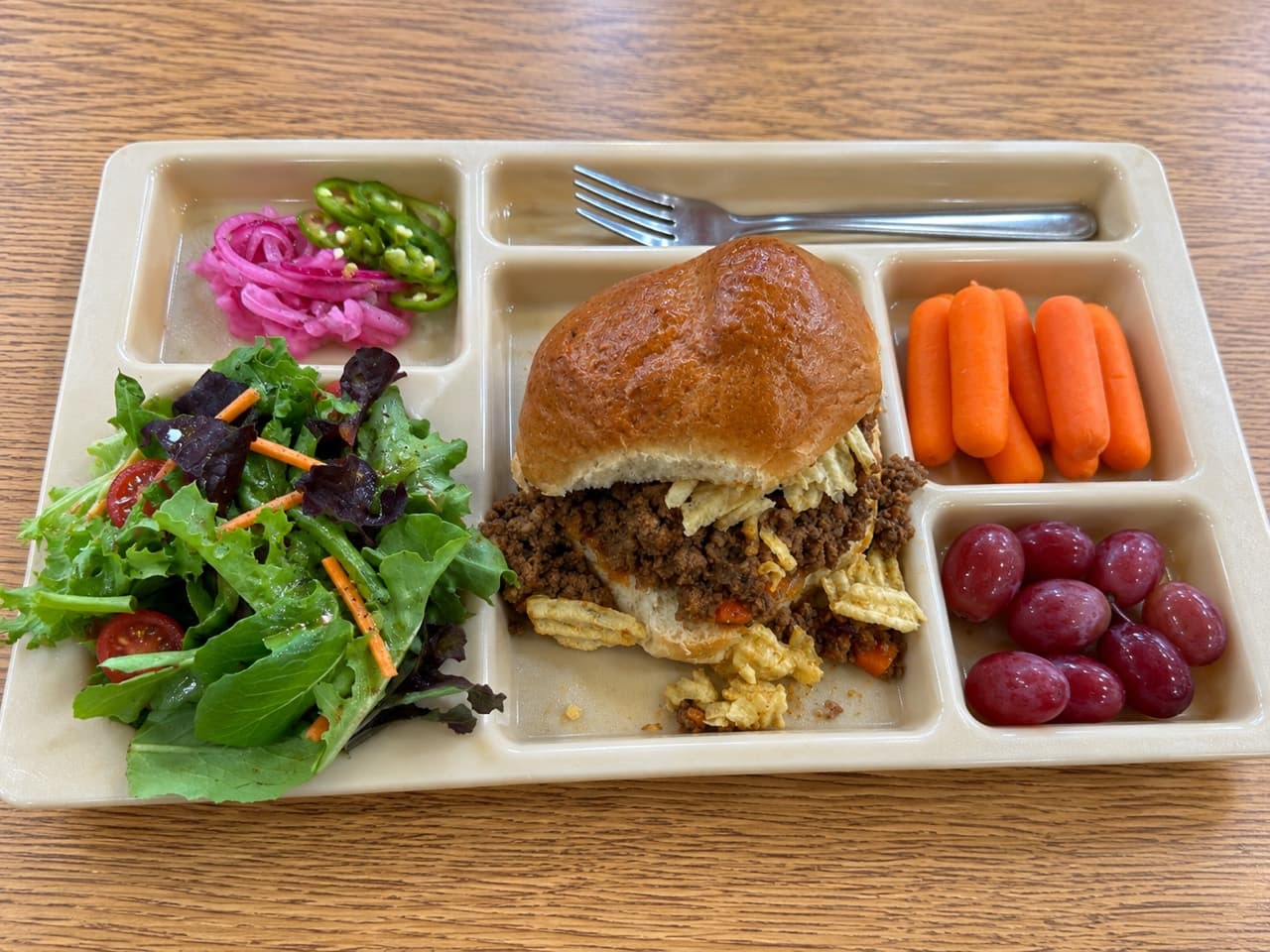 A Wagyu sloppy joe topped with crushed kettle chips on a school lunch tray, with salad, carrots, grapes and pickled vegetables.
