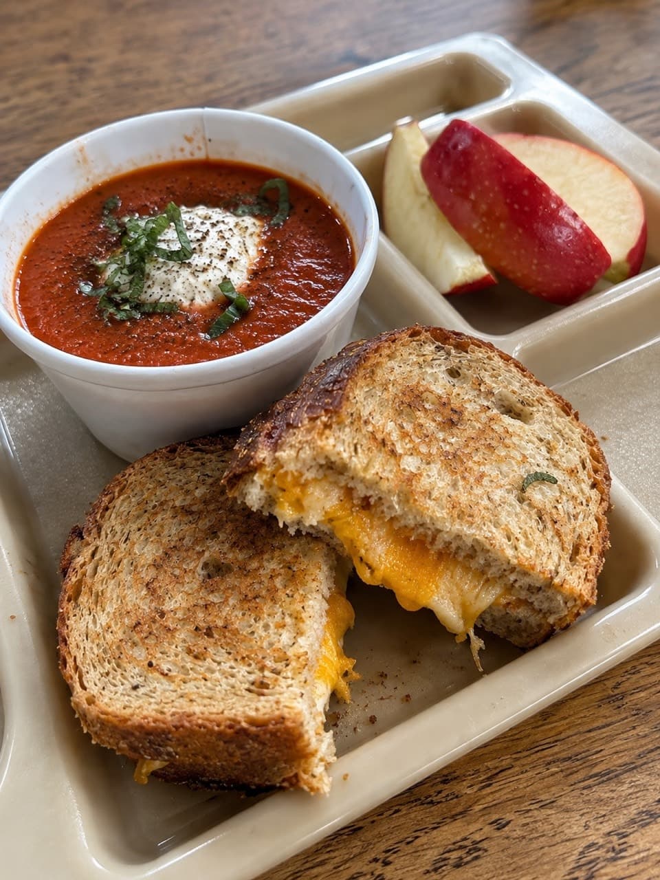 Tomato soup topped with herbs, two golden grilled-cheese halves and sliced red apples on a lunch tray.