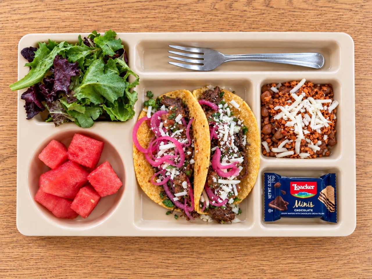 Guajillo barbacoa tacos topped with pickled red onions, cheese and herbs on a lunch tray.